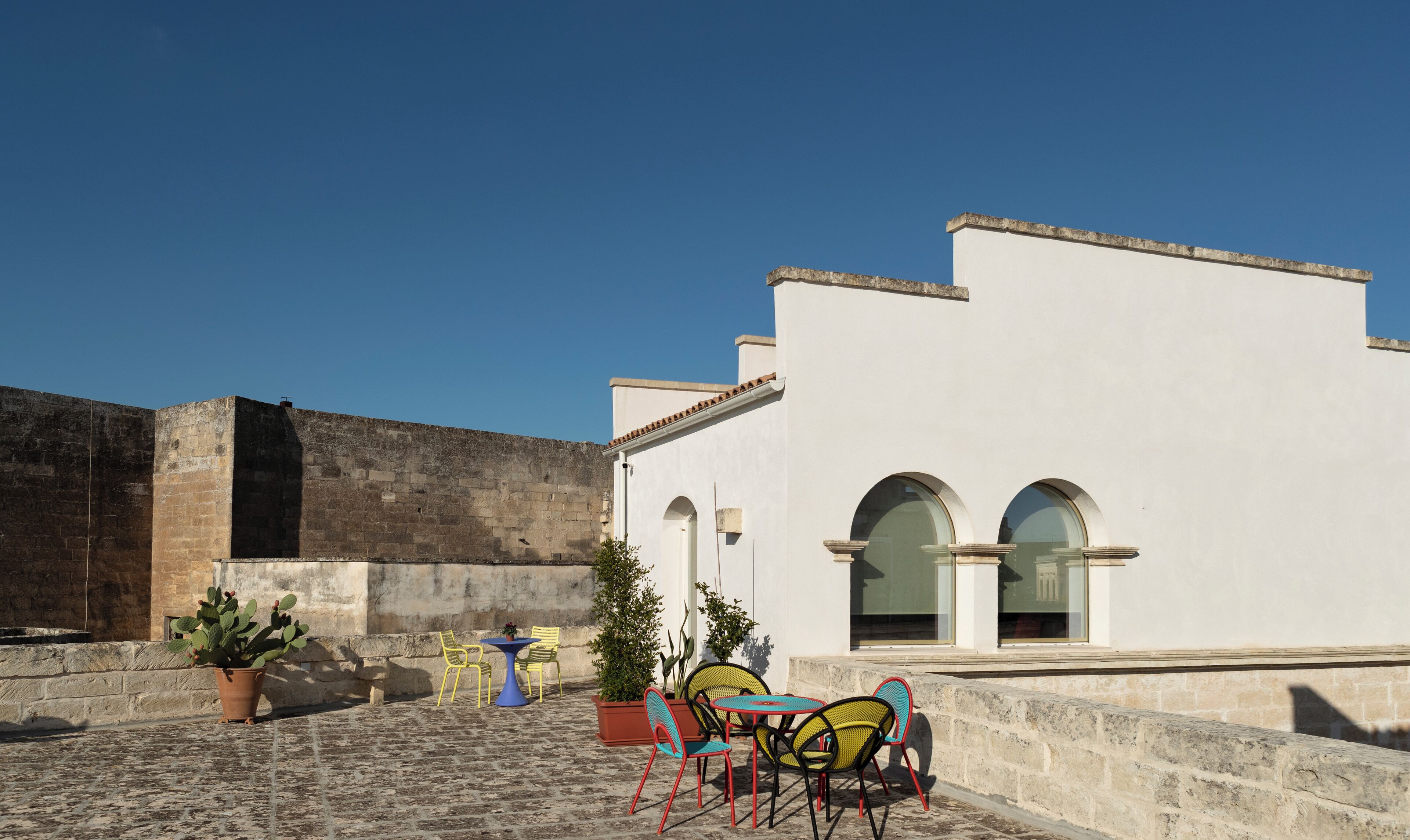 A courtyard with a white building featuring arched windows and a stone wall. There are colorful chairs and a table with potted plants.