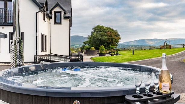 A hot tub with a bottle of champagne and glasses, a wooden deck, and a view of a house and mountains.