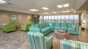 Amplia sala de estar con un sofá modular grande, varios sillones y una mesa de centro. Se observa una estantería llena de libros y objetos decorativos. La habitación cuenta con grandes ventanales que ofrecen vista al océano.