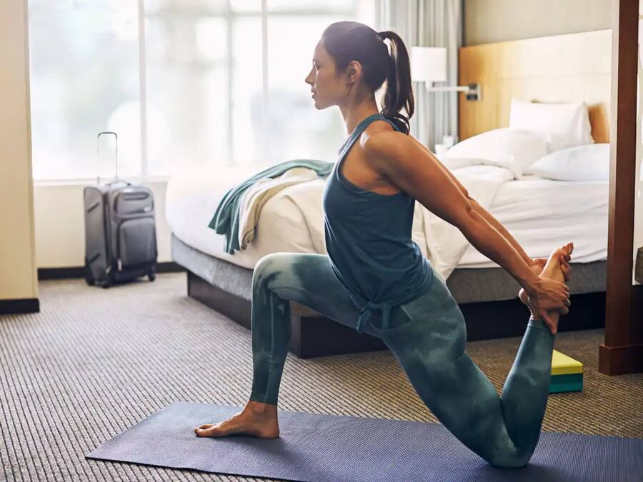 Una mujer realizando una postura de yoga sobre una colchoneta en una habitación de hotel, con una cama y una maleta al fondo.