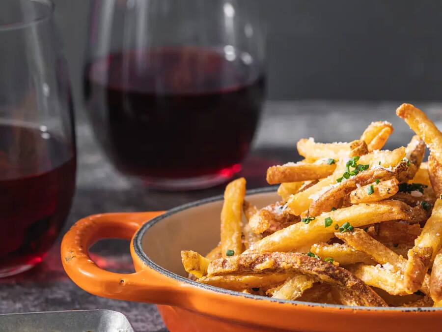 Un plat de patates fregides cruixents en un bol, acompanyat d'un bol petit de salsa i un got de vi vermell.
