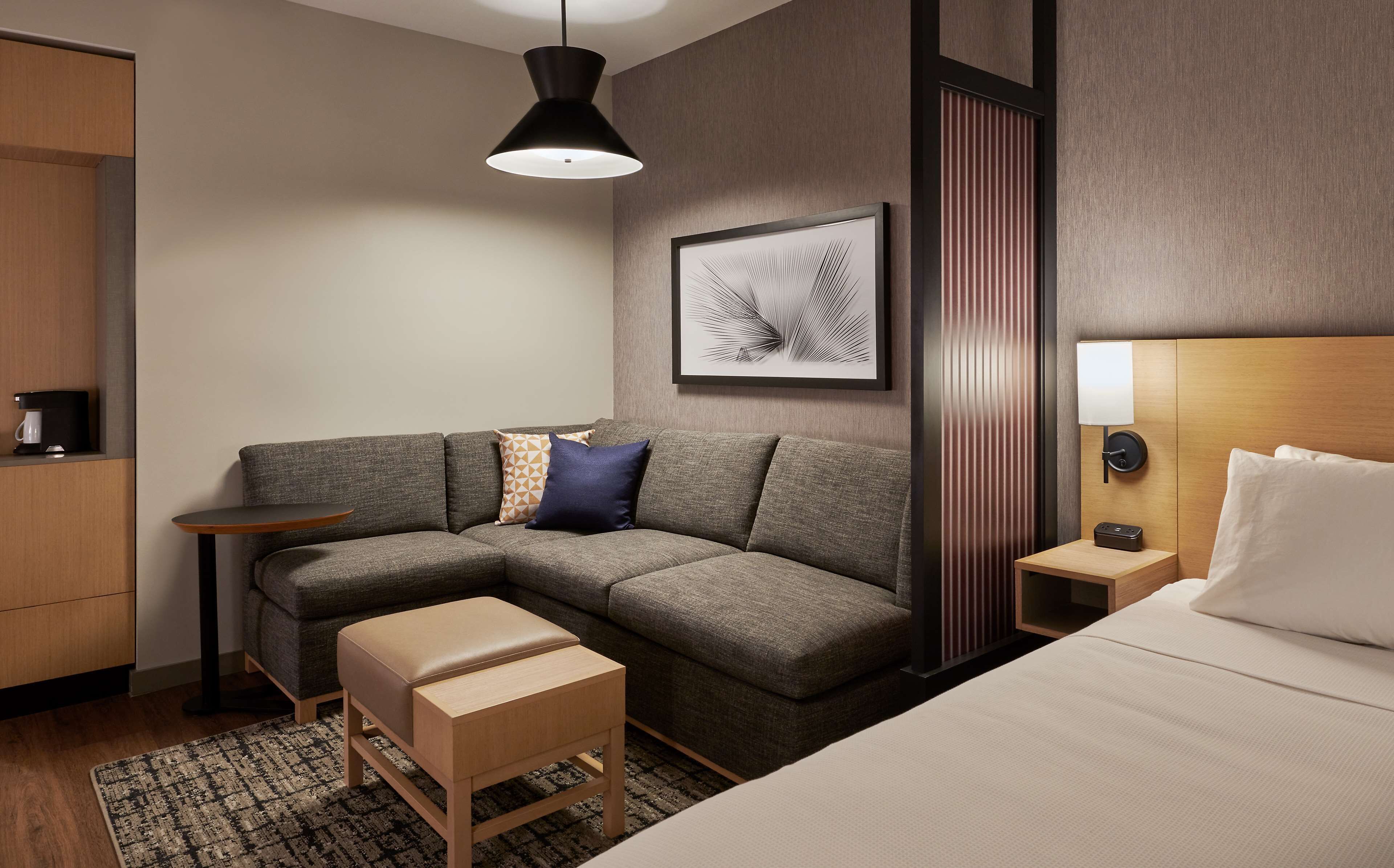 Ein modernes Hotelzimmer mit einem grauen Ecksofa, einer Holzbank, einem kleinen Beistelltisch, einer Wandleuchte und einem gerahmten Kunstwerk.