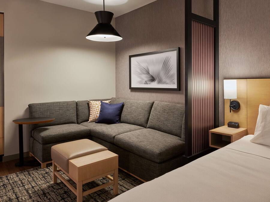 Quarto de hotel moderno com sofá modular cinza, um banco de madeira, uma mesinha de apoio, luminária de parede e uma obra de arte emoldurada.