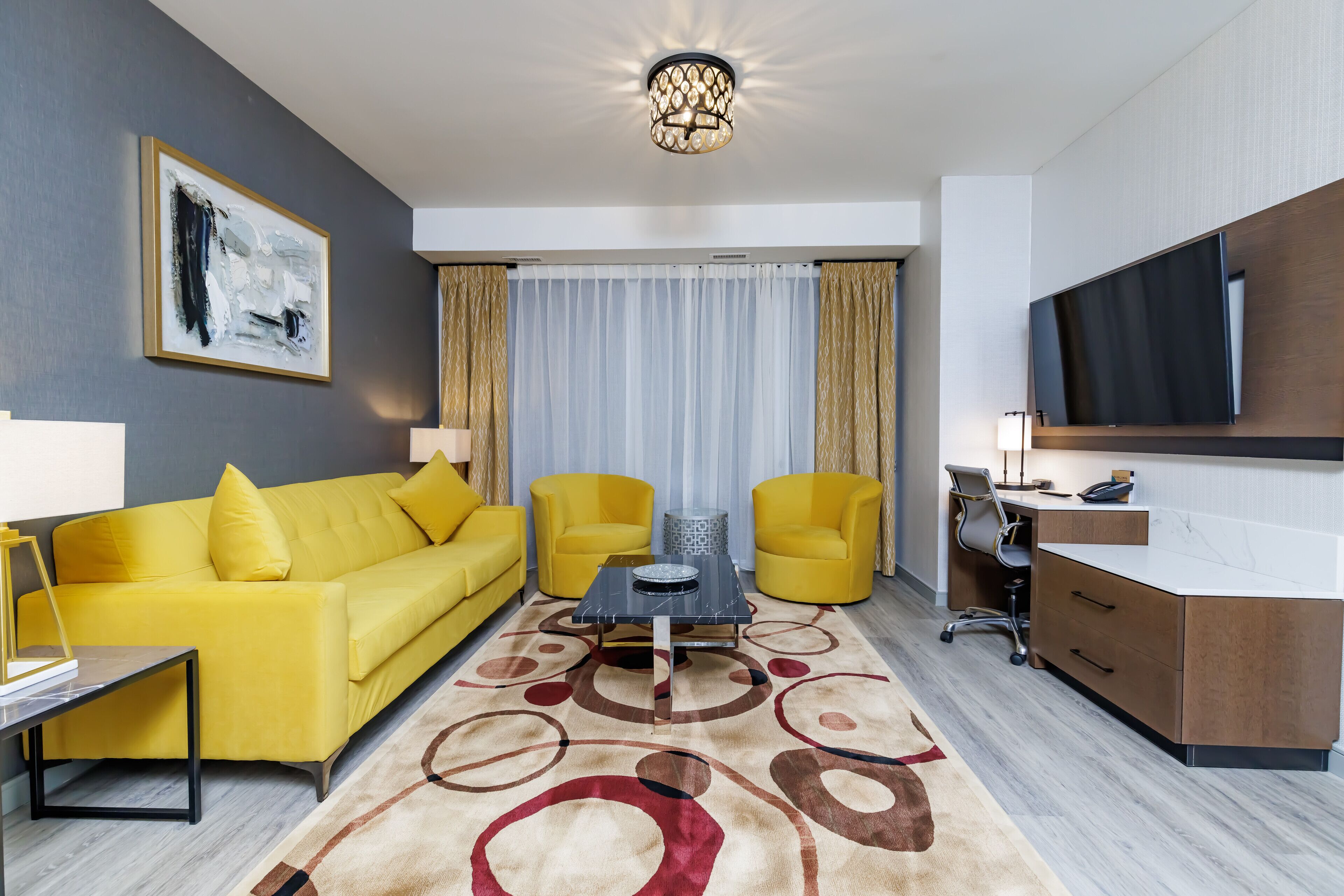 Quarto de hotel moderno com sofá amarelo, uma mesa de centro, escrivaninha com cadeira, televisão de tela plana e uma grande janela com cortinas.