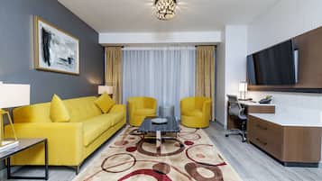 Quarto de hotel moderno com sofá amarelo, uma mesa de centro, escrivaninha com cadeira, televisão de tela plana e uma grande janela com cortinas.