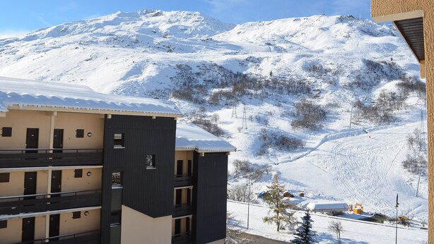 A multi-story building with balconies, a snow-covered mountain in the background, and a ski slope with tracks. - LES MENUIRES apartment sleeps 8 2 bedrooms + mezzanine (LES BELLEVILLE)