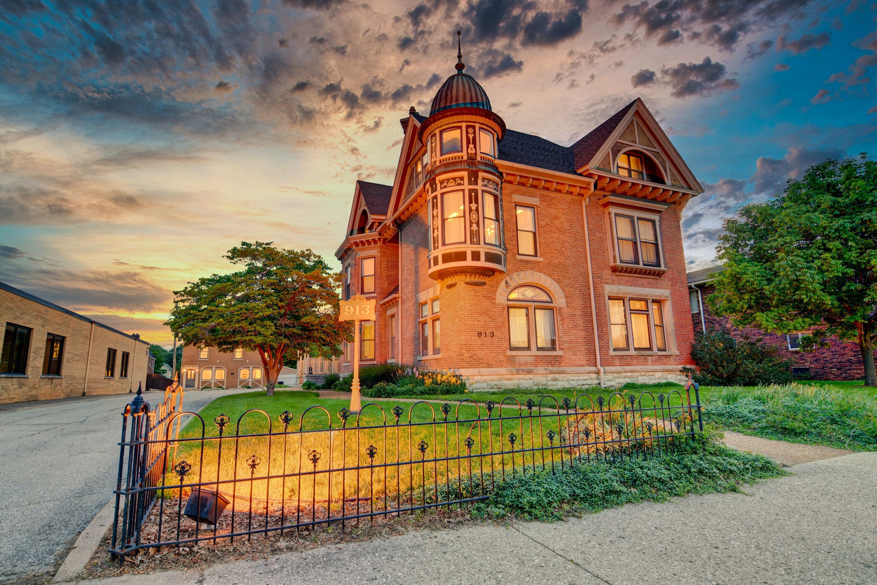 A large, historic brick building with a turret and multiple windows, surrounded by a well-maintained lawn and a metal fence.