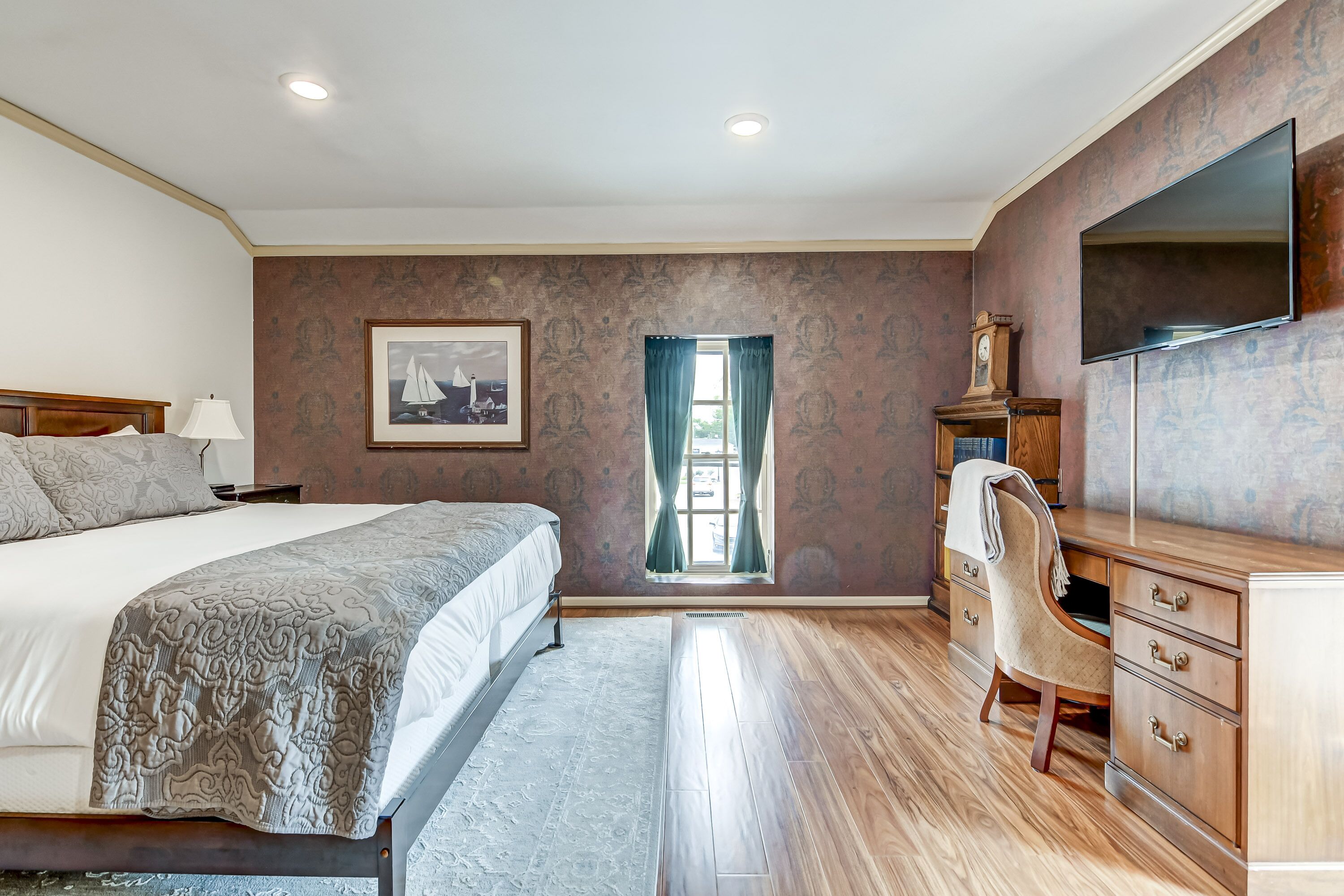 Een slaapkamer met een bed, bureau, stoel, televisie, raam met gordijnen, en een ingelijste foto aan de muur.