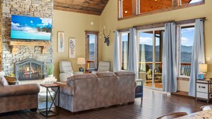 A spacious living room with a stone fireplace, comfortable seating, a flat-screen TV, and large windows offering a view of the mountains. - Star Of The Summit, Private Pool, Breathtaking Mountain Views, FREE Tickets! (Sevierville)