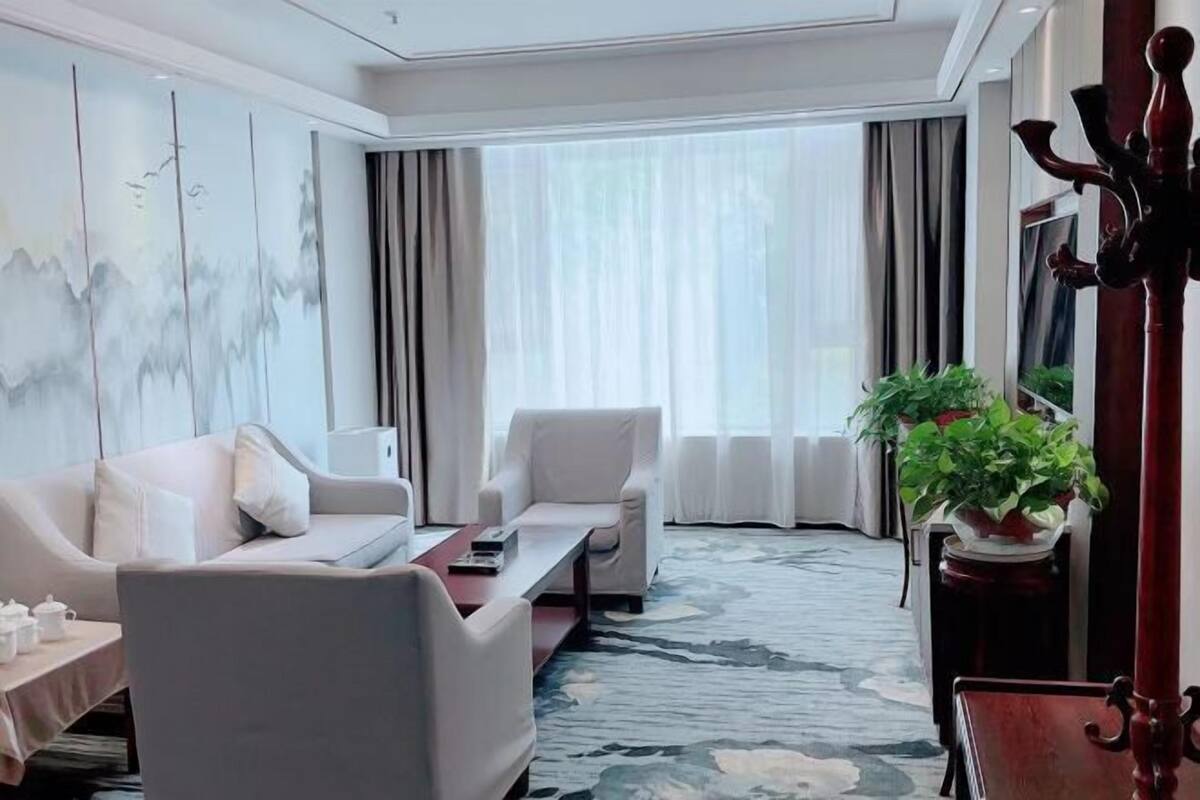 A modern hotel room with a sofa, armchair, coffee table, and a large window with curtains.