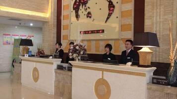 A hotel reception desk with two receptionists, a digital clock, and a decorative wall with a map.