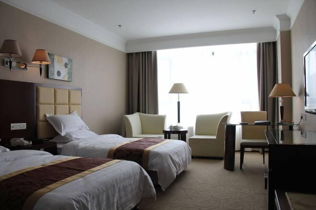 A hotel room with two beds, a desk, a chair, a lamp, and a large window.