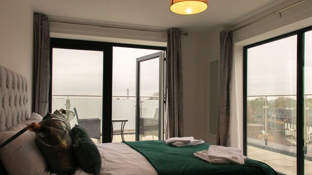 Een hotelkamer met een bed, een groene plaid, een raam met uitzicht op de stad en een balkon met een stoel.
