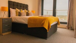 Een slaapkamer met een bed, nachtlampjes, een nachtkastje en uitzicht door een groot raam.
