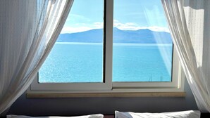 A window with white curtains, a bed with white pillows, and a view of a calm sea and mountains. - ERAL Apart-Hotel (Sarandë)