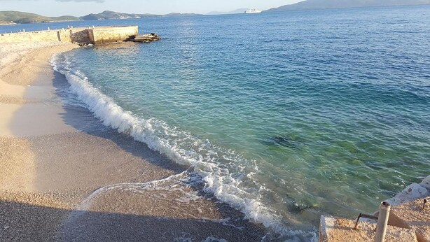 A beach with clear water, a stone wall, and a distant ship. - ERAL Apart-Hotel (Sarandë)