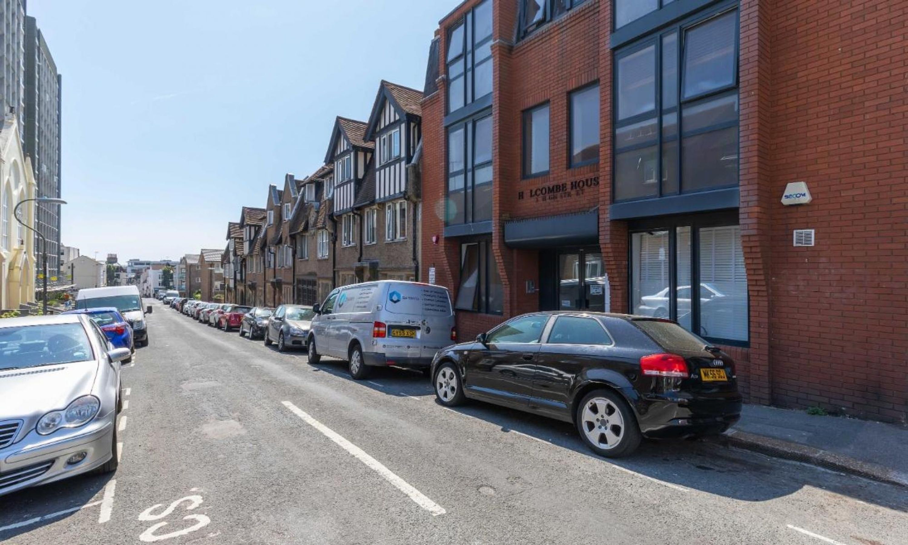 A row of parked cars along a street with a mix of residential and commercial buildings.