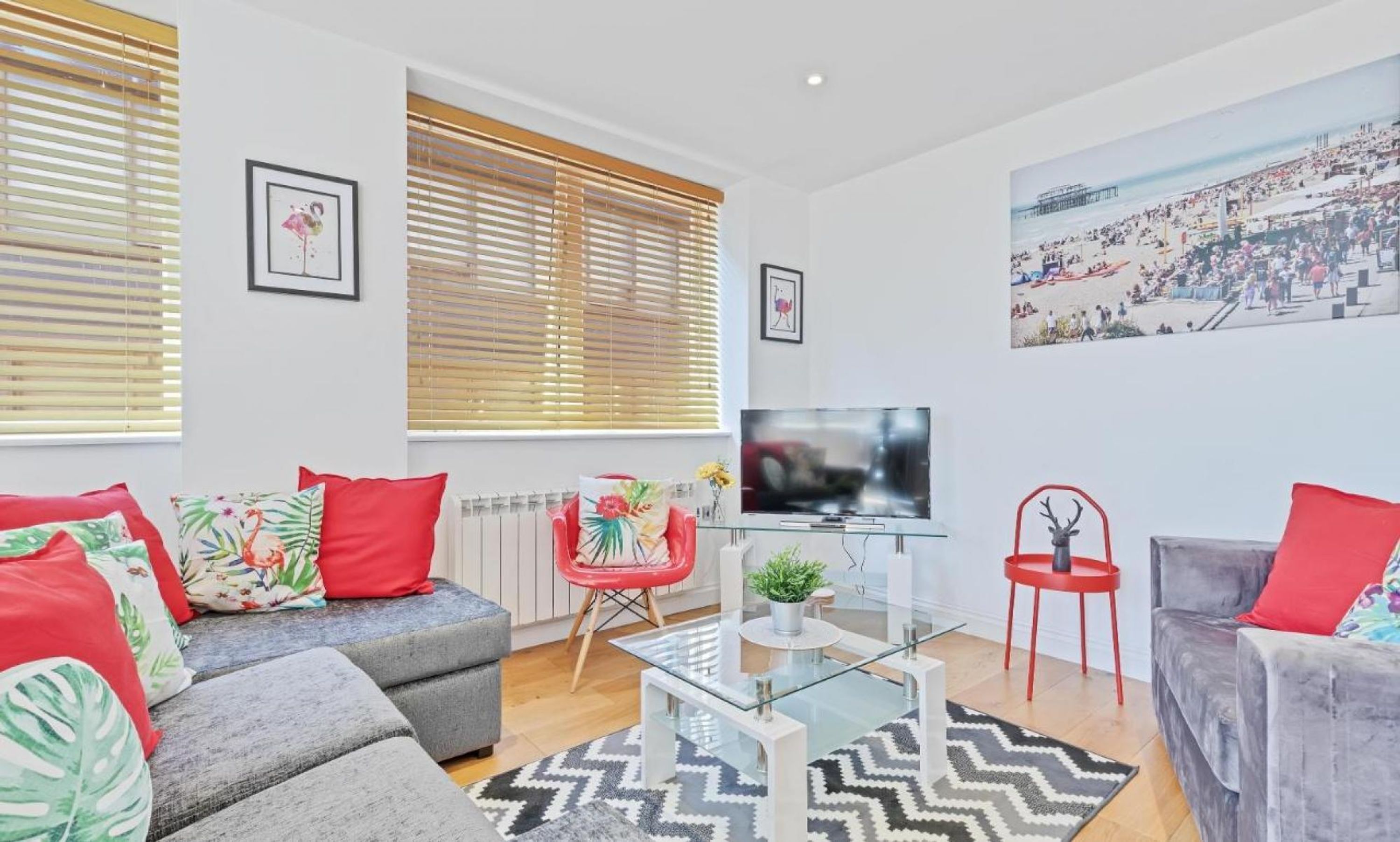 A modern living room with a grey sofa, red and floral cushions, a glass coffee table, and a TV.