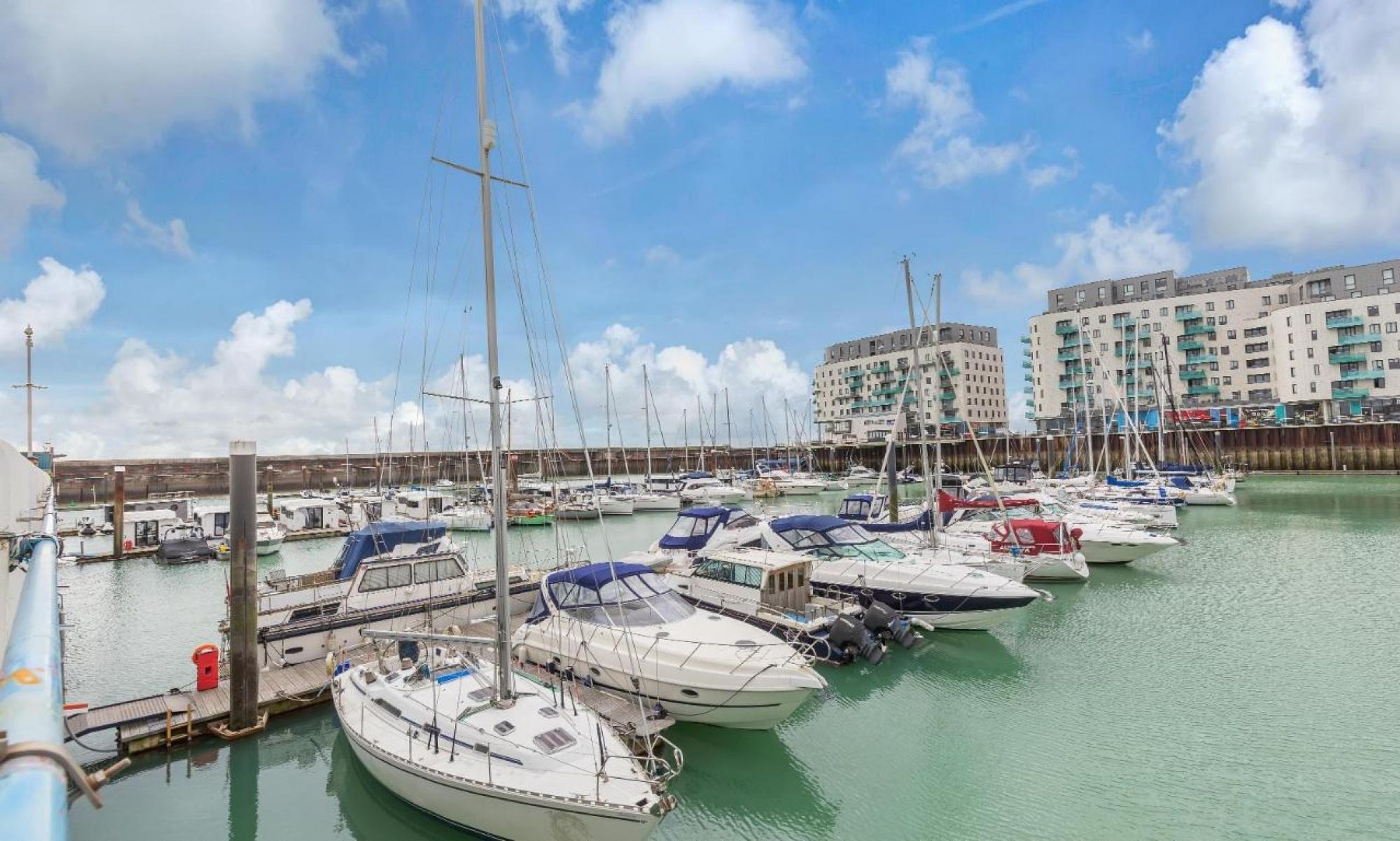 A marina with multiple boats docked, a pier, and buildings in the background.