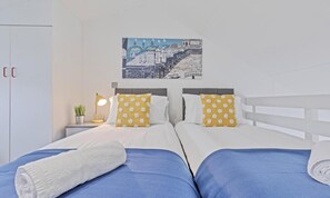 Two single beds with white bedding and a yellow pillow, complemented by a potted plant and a cityscape photograph.