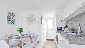 Cozinha moderna com armários brancos, pia de aço inoxidável e bancada. Há uma área de refeições com uma mesa posta para dois, cadeiras brancas e um vaso de planta.