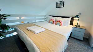 Uma cama bem arrumada com edredom branco e duas almofadas com padrão de arco-íris, uma mesa de cabeceira com abajur e um quadro na parede.