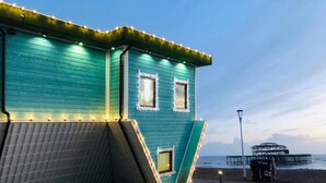 A teal-colored building with two windows and a slanted roof, situated by a beach with a pier in the background.