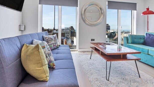 A modern living room with a grey sofa, a blue sofa, a wooden coffee table, and a city view through large windows.