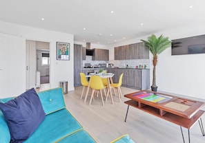 A modern living room with a teal sofa, a wooden coffee table, and a dining area with yellow chairs. - Right On Bright On Apartment 1B - City Centre Luxury Apartments (Brighton)
