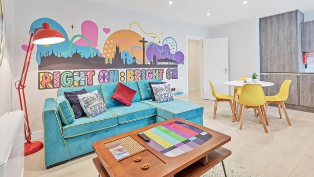 A modern living room with a teal sofa, a wooden coffee table, and a colorful wall mural. - Right On Bright On Apartment 1G - City Centre Luxury Apartments (Brighton)