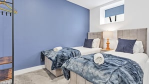 A hotel room with two beds, a blue wall, a wooden ladder, and a window with blinds. - Right On Bright On Apartment 1G - City Centre Luxury Apartments (Brighton)