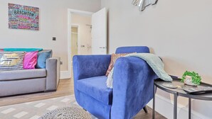 Un salon avec un fauteuil bleu, un canapé gris et une petite table ronde sur laquelle se trouvent une plante et une télécommande.