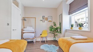 Une chambre d’hôtel avec deux lits, une armoire en bois, une chaise jaune, une plante en pot et une fenêtre donnant sur la rue.