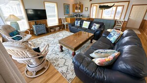 Een woonkamer met een zwarte leren bank, een houten salontafel, rieten fauteuils en een televisie.