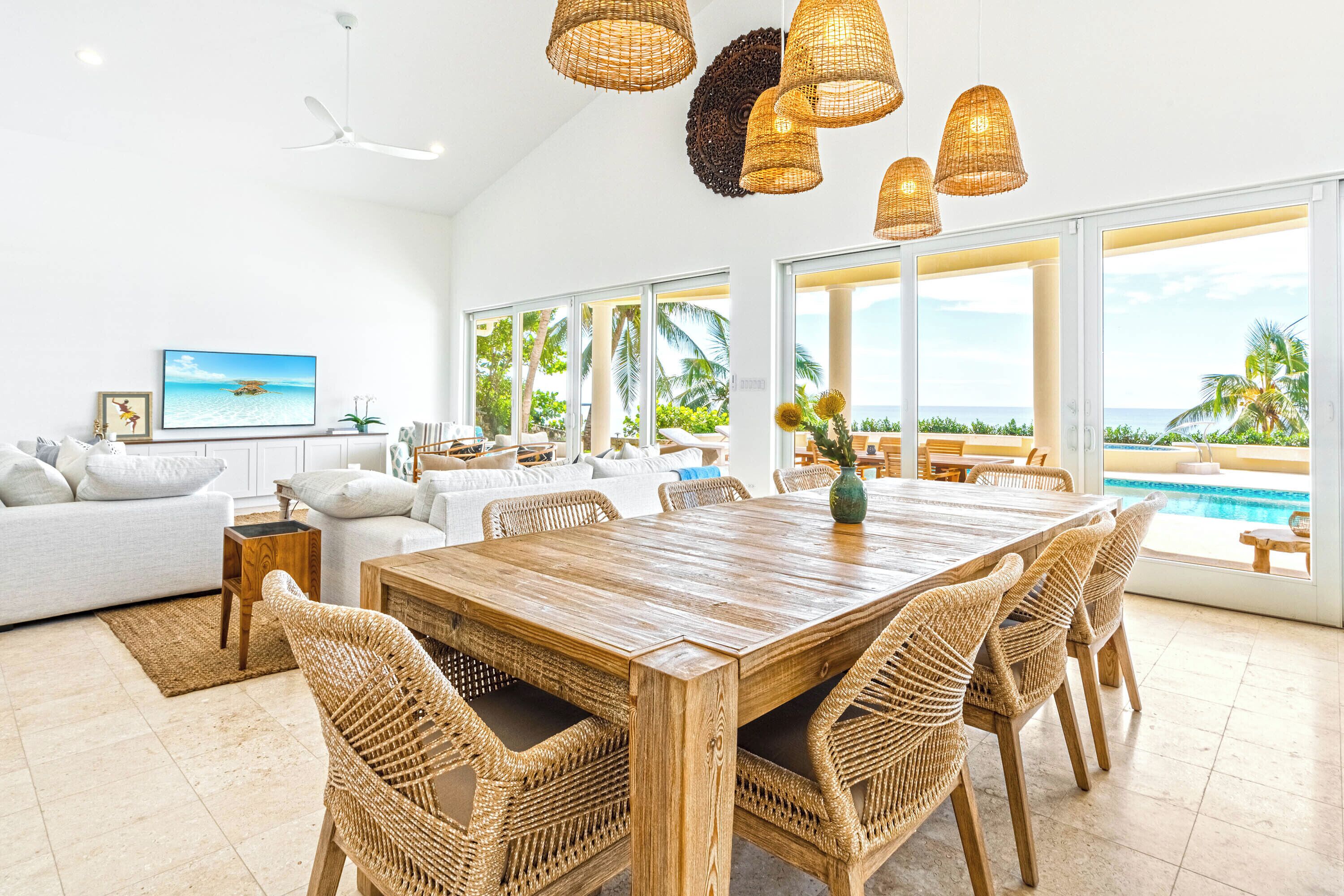 A spacious living area with a large wooden dining table surrounded by wicker chairs, a white sofa, and a television on the wall.
