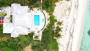 Ett flygbildsfoto av en fastighet vid stranden med en swimmingpool, palmer och en vit byggnad med en unik takdesign.