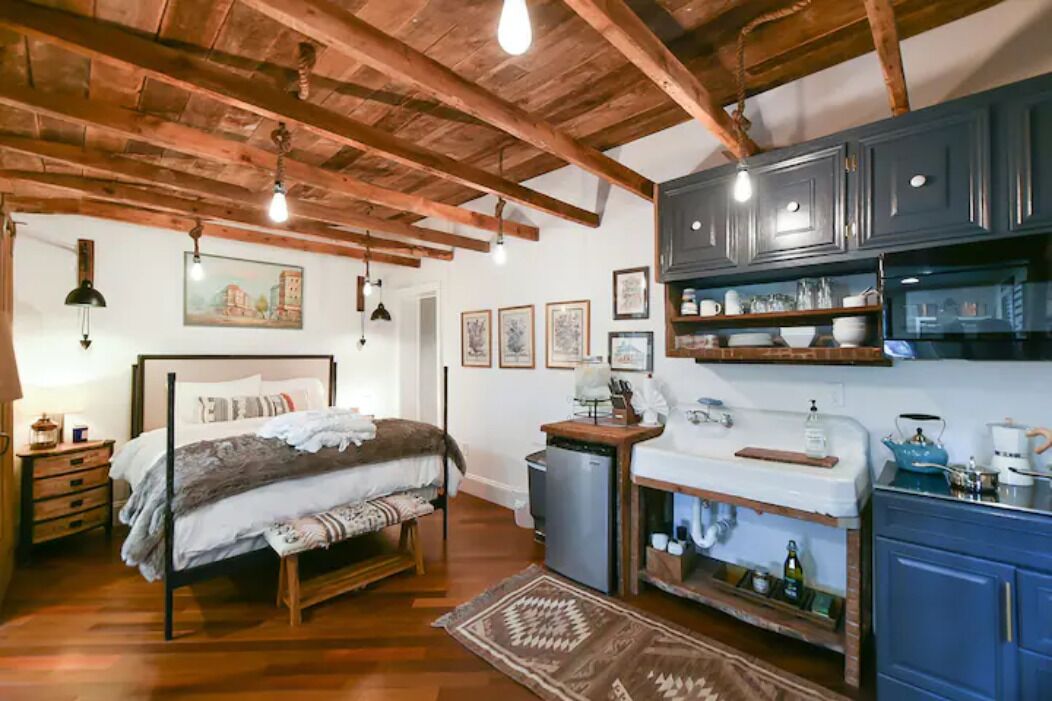 Een slaapkamer met een houten bed, een klein bureau en een kitchenette met blauwe kastjes.