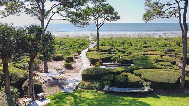 Un jardín ajardinado con un sendero que lleva a la playa, rodeado de setos cuidados y palmeras.