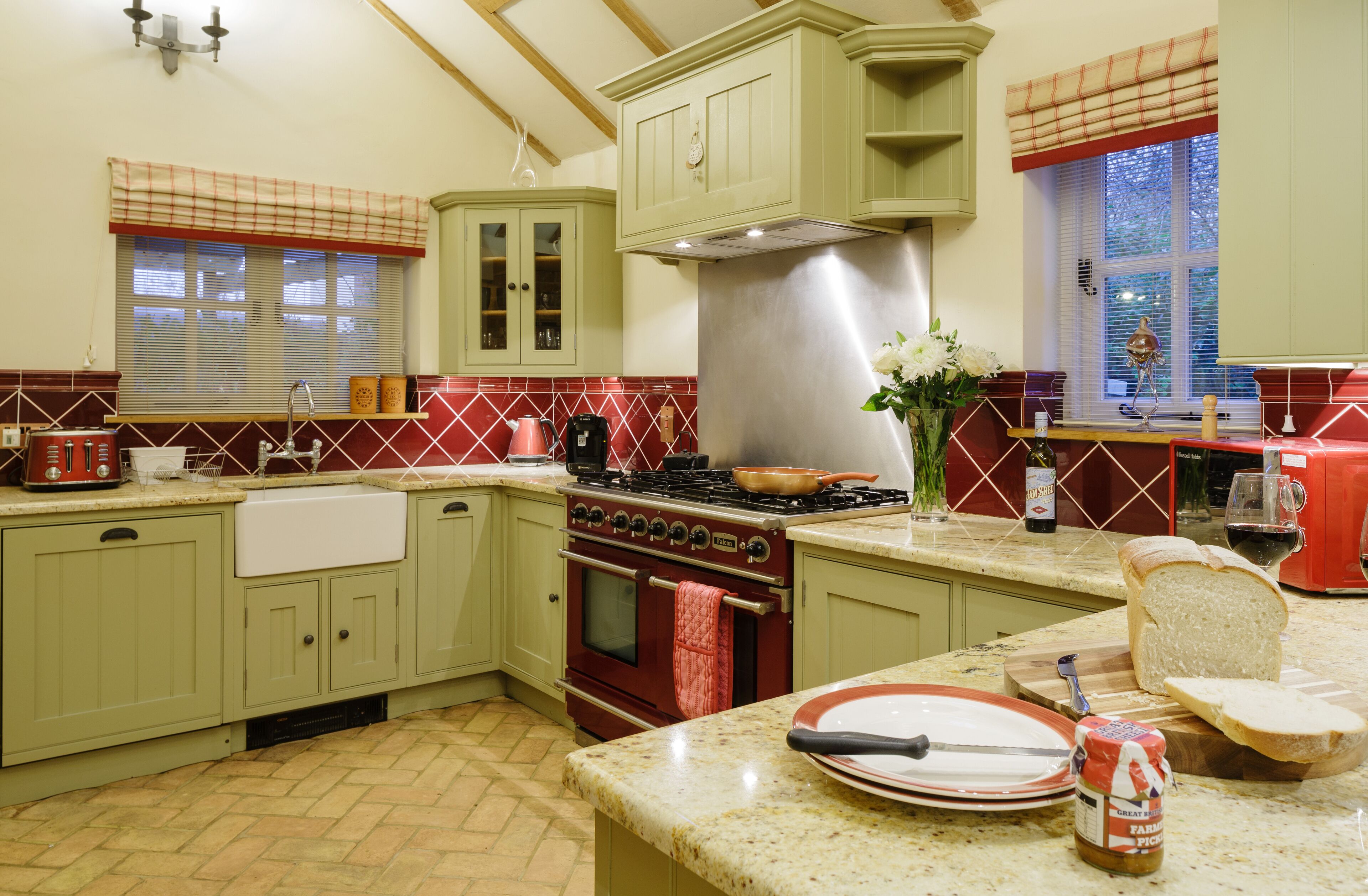 Una cocina con gabinetes verdes, una campana extractora de acero inoxidable, un salpicadero de azulejos rojos y una ventana con persianas.