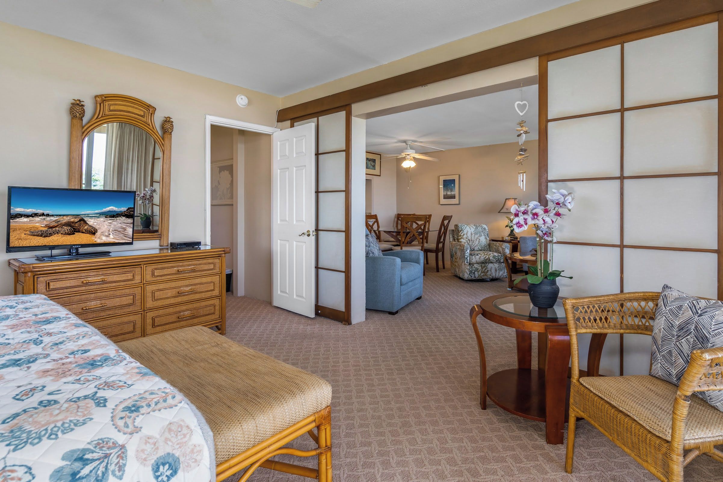 A hotel room with a bed, a television, a dresser, a dining area with chairs, and a view of the beach through a large window.