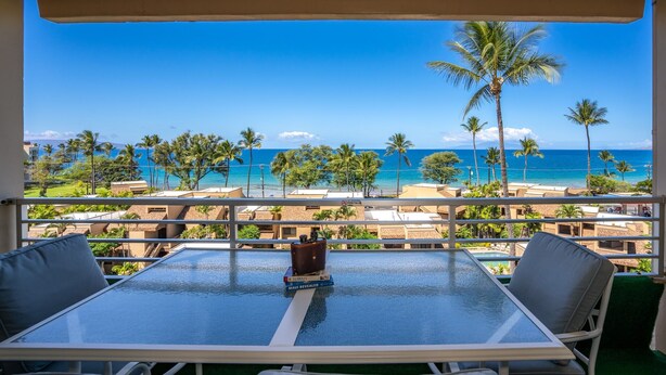 A balcony with a glass table, two chairs, and a view of the beach and ocean.