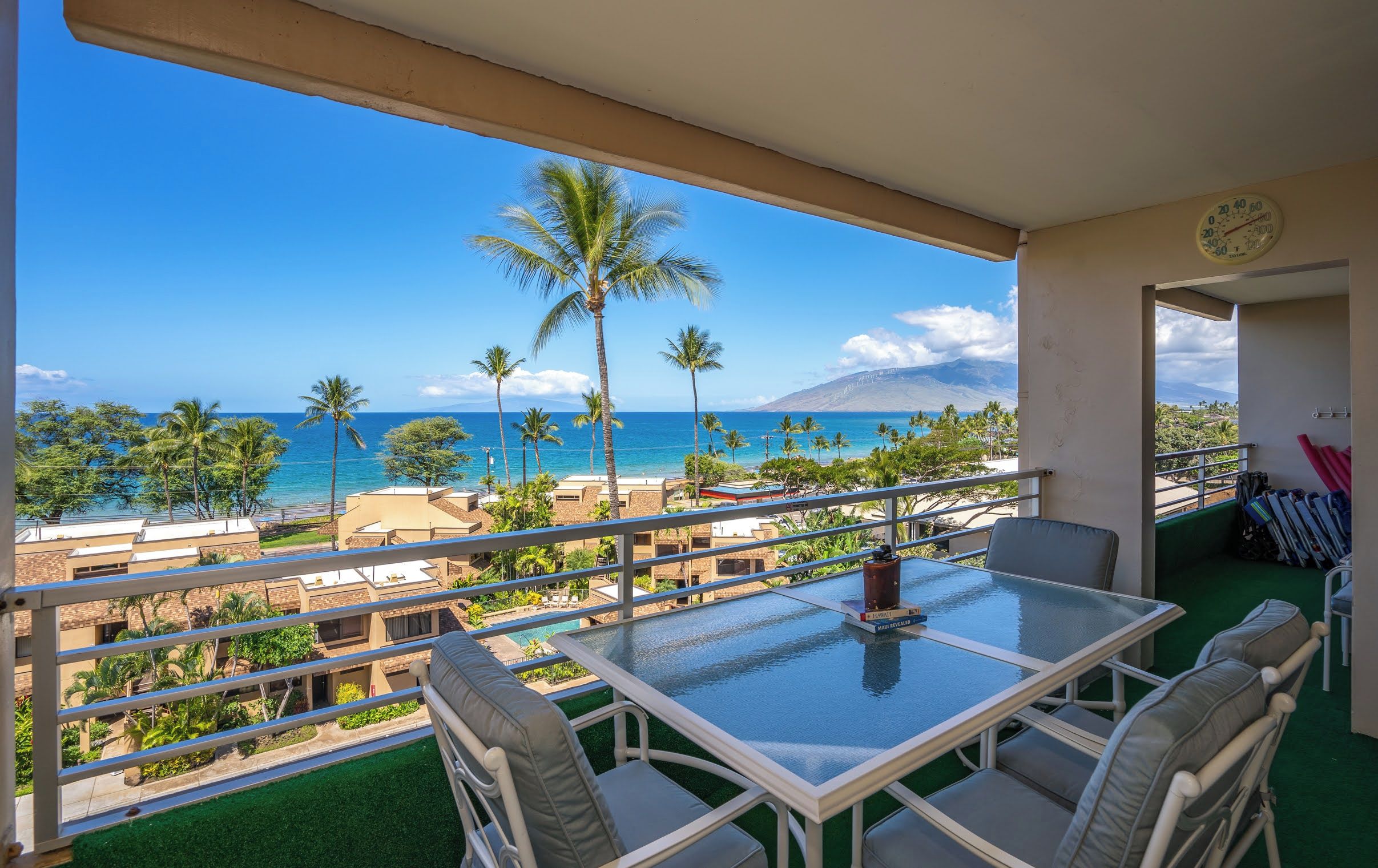A balcony with a glass table and chairs, overlooking a beach with palm trees and a clear blue sky.