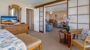 A hotel room with a bed, a television, a dresser, a dining area with chairs, and a view of the beach through a large window.