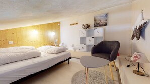 Un dormitorio con cabecera de madera, cama con ropa de cama blanca, un sillón gris, una mesita redonda pequeña, una lámpara y una estantería con objetos decorativos.