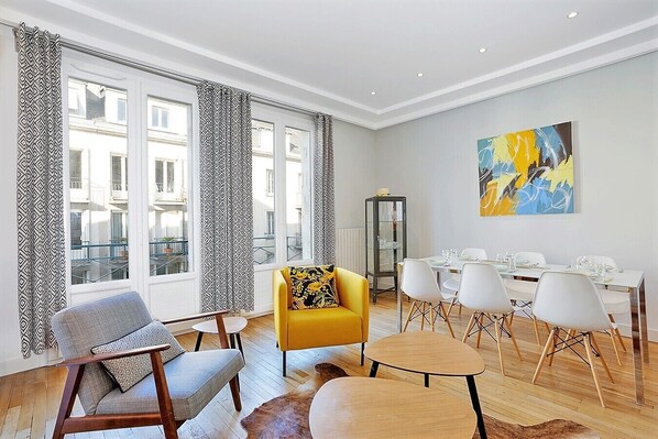 A modern living room with a yellow armchair, a grey armchair, and a dining area with white chairs and a glass cabinet.