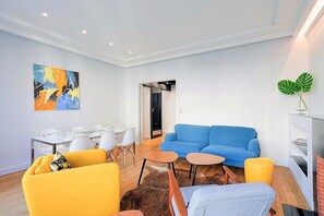 Ein modernes Wohnzimmer mit einem blauen Sofa, einem gelben Sessel und einem Essbereich mit weißen Stühlen und einem Tisch.