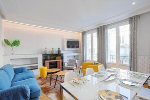 A modern living room with a sofa, a dining table set for four, a fireplace, and large windows with patterned curtains.