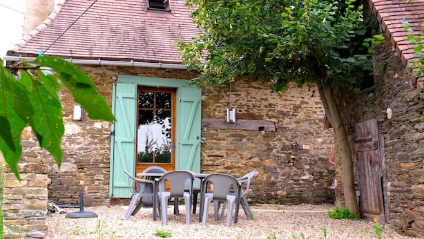 A rustic stone building with a tiled roof, a wooden door with green shutters, and a small outdoor seating area with plastic chairs.