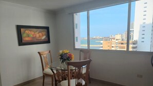 Un coin repas avec une table en verre et des chaises en bois, un tableau encadré au mur, et une vue sur le paysage urbain et l’eau, visible à travers de grandes fenêtres.