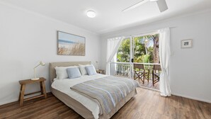 A bedroom with a bed, a nightstand, a desk, a chair, a balcony with a view of greenery, and a ceiling fan.